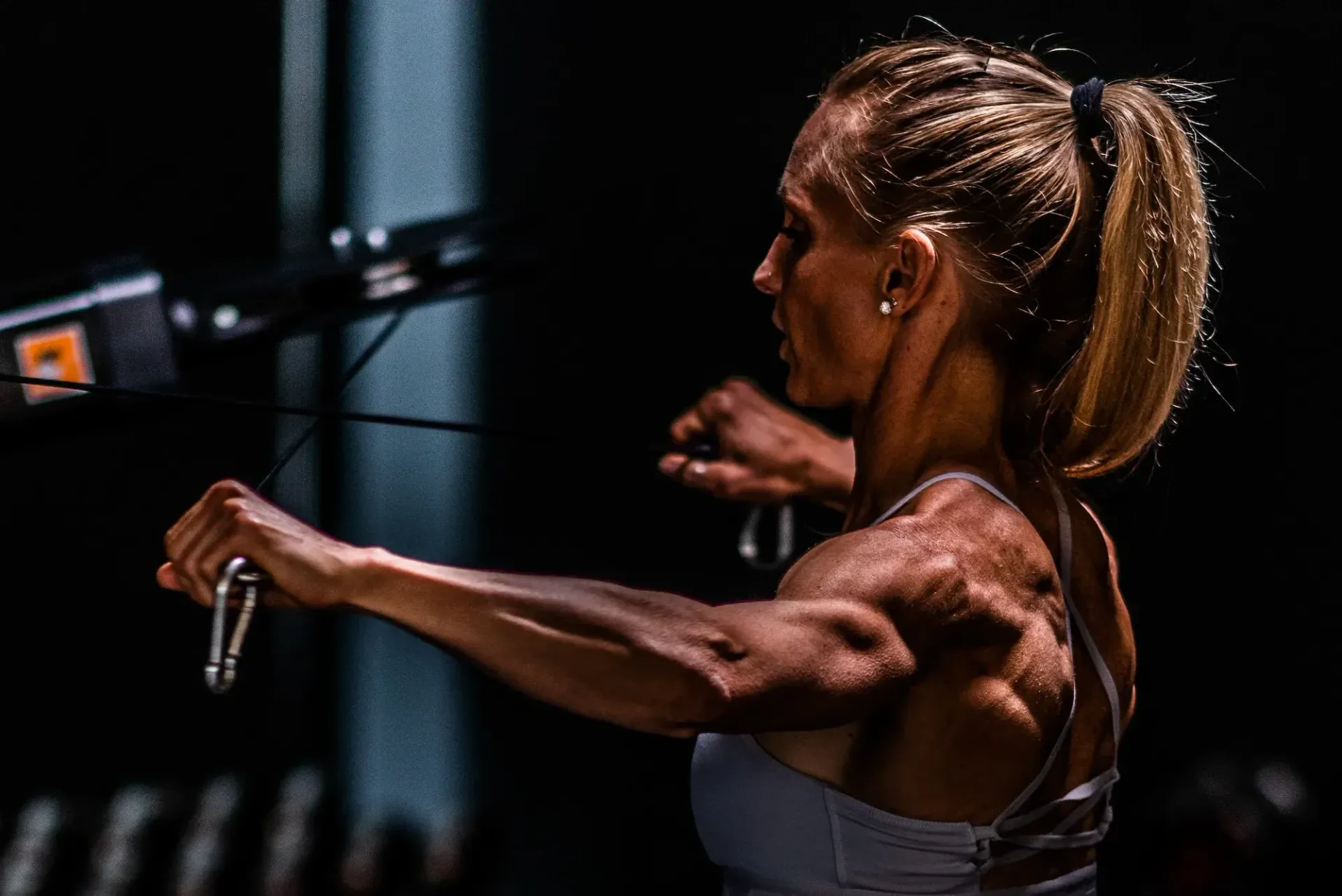 Blonde fitness trainer in black outfit coaching a woman on a cable machine in a bright gym.